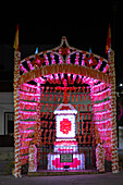 Patron saint celebrations light up Castilleja del Campo with vibrant decorations and a cross adorned with colorful paper at night.