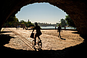 People explore cobblestone paths near Muelle de la Sal beneath a bridge in Seville.