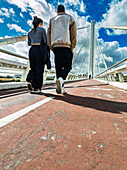 A young couple enjoys a leisurely walk on the bike and pedestrian lane of Alamillo Bridge in Seville, Andalusia, under a blue sky.