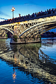 Worshippers admire El Cristo del Cachorro as it shines above Triana Bridge, creating a stunning reflection in the river at dusk.