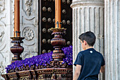 A young boy leans against columns to observe a procession in Seville, Spain.