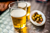 Glass of beer served alongside spiced olives on a plate against a blurred background.