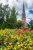 Vibrant flowers bloom in the foreground as Paulus Church's bell tower rises against a cloudy Oslo sky.