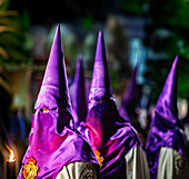 Hooded penitents from the Cigarreras brotherhood participate in a solemn procession, holding candles in the vibrant streets of Seville.