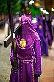 Penitents in purple robes participate in a solemn procession, bearing wooden crosses, celebrating Holy Week in Seville, Andalusia.