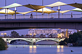 Cyclists and kayakers enjoy the evening by the Cristo de la Expiracion Bridge in Seville, with the Triana Bridge glowing nearby.