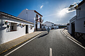 A person walks along a sunlit street in El Madroño, surrounded by whitewashed buildings adorned with flowers during a clear day.