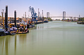Angedockte Schiffe im Hafen von Sevilla mit Kränen im Vordergrund und der Centenario-Brücke, die beide Flussufer bei klarem Himmel verbindet.