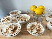 Schalen mit cremigem, mit Zimt bestreutem Dessert stehen neben frischen Zitronen auf einer hölzernen Oberfläche und bilden eine helle, einladende Auslage.