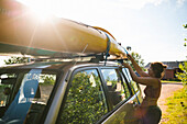 Seitenansicht einer Frau, die an einem sonnigen Tag im Urlaub ein Kajak auf die Motorhaube eines Autos setzt