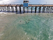 Aerial view of surfers riding waves near a bustling pier and tranquil beach, Oceanside, California, United States.