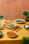 Ein üppiges italienisches Abendessen mit Schüsseln mit Basilikum-Pesto-Gnocchi, frischem grünen Salat, Süßkartoffelstückchen und cremigem Ricotta-Käse. Die Szene ist auf einer leuchtend gelben Tischdecke vor einem orangefarbenen Hintergrund mit grünen Blättern angerichtet.