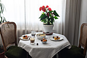 Warmly lit coffee table covered in a white cloth with various desserts such as cherry cake and ice cream, a red flowering potted plant, coffee and refreshing water, creating an inviting and peaceful atmosphere.