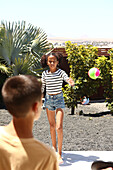 A young girl and a boy are playing tag in a sunny backyard garden. The girl in a striped top and jeans shorts prepares to throw a colourful ball. Lush greenery surrounds her.