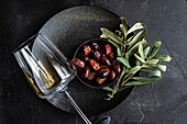 Top view of a sophisticated arrangement of Greek BBQ olives in a black bowl with fresh bay leaves, accompanied by a glass of dry white wine, all set against a dark, textured background.