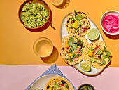 Viewed from above, the ingredients for tacos are presented on a yellow and pink background. On the plate are tacos with mango, coriander, avocado, lime and spiced fish, accompanied by guacamole, pickled onions and a glass of drink.