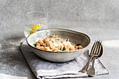A ceramic bowl of cavatappi pasta, topped with cheese and savoury tomato sauce, served on a napkin, alongside cutlery and a refreshing lemon water on a textured surface.