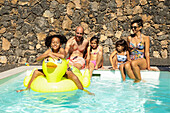 Family relaxing by the pool, a young girl on a yellow rubber ducky. Parents and children in swimming costumes enjoy the sunny day on the stone wall, splashing around and smiling.
