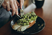 Eine Person schneidet Avocado-Toast mit Kernen auf einen dunklen Teller. Der Fokus liegt auf dem Esserlebnis, der Hintergrund ist unscharf und unterstreicht die warme und gemütliche Atmosphäre.
