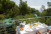 Ein Hotelbalkon mit Blick auf eine sonnenbeschienene türkisfarbene Bucht, umrahmt von üppigen grünen Bäumen Ein elegant gedeckter Frühstückstisch mit frischem Obst, Gebäck und Getränken sorgt für einen perfekten Morgen