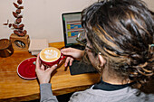 A person holds a latte with art in a red cup and works at a wooden desk with a laptop. The scene combines productivity with the enjoyment of a speciality coffee.