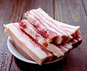 Slices of raw pork belly are neatly arranged on a white plate on a rustic wooden platter. The marbled fat and rich red meat emphasise its freshness.