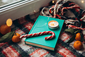 Cosy Christmas ambience with a burning candle on a turquoise-coloured book, surrounded by a candy cane, clementines and a plush blanket by the window.
