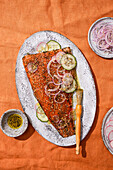 A delicious roasted salmon fillet, flavoured with herbs and sesame seeds, garnished with sliced onions and cucumber on a rustic plate. Next to it is a small bowl of olive oil.