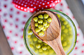 A wooden spoon holds green, home-grown organic peas over a glass jar resting on a white cloth with red polka dots. The bright colours and textures create a fresh, inviting scene.