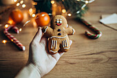 A hand holds a gingerbread man decorated with icing In the background, blurred festive elements such as candy canes and oranges create a cosy holiday atmosphere