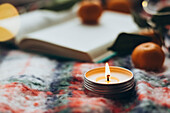 A burning candle on a colourful blanket, accompanied by a book and oranges that create a warming Christmas atmosphere.