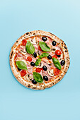 Top view of a delicious mozzarella pizza topped with bacon, cherry tomatoes, black olives, basil and mushrooms, served on a bright turquoise background.