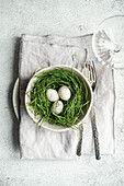 Top view of a simple but elegant Easter table setting with a bowl containing a nest of fresh green grass and three speckled eggs. A pair of vintage forks completes the cheerful tableau, which is set on a soft linen napkin against a light-coloured, textured background.