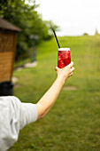 A hand holding a refreshing red drink with ice and a straw against a lush green background on a bright, sunny day that creates a perfect summer atmosphere