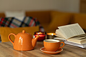 A cosy setting with an orange teapot and cup on a wooden table. An open book and lit candles create a warm, inviting atmosphere that invites you to relax and read.