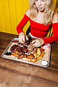 A woman in a red top enjoys a barbecue with ribs, sausages and various types of meat presented on a tray. The wooden table and the bright yellow background emphasise the scene.