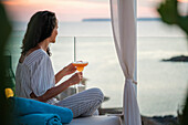 A woman relaxes on a terrace and sips a cocktail at sunset. The tranquil setting overlooking the sea provides a peaceful and picturesque backdrop for leisure.