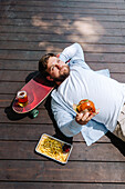 A person enjoys a relaxing moment on a wooden terrace with a burger in their hand. Nearby is a skateboard, fries and a drink, capturing the essence of a relaxed barbecue day.