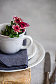 A rustic white ceramic cup filled with bright pink flowers stands on a wooden surface A soft grey napkin lies underneath, creating a cosy, inviting atmosphere