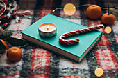 Cosy Christmas atmosphere with a burning candle on a blue-green book, candy canes and oranges, surrounded by festive decorations.