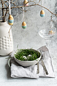 An elegantly laid Easter table with a bowl full of eggs in fresh green grass, accompanied by a delicate arrangement of hanging, artistically painted eggs and a sophisticated ceramic vase.
