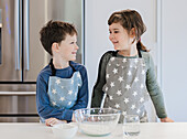 Two children in matching aprons with a star pattern stand at a kitchen table and smile at each other. A bowl of ingredients and a glass of water stand next to them.