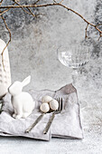 Ein elegantes Osterthema mit einem weißen Keramikhasen, drei gesprenkelten Eiern und einem klaren Glas. Ergänzt wird dieses Arrangement durch Silberbesteck und eine strukturierte Serviette, die für eine heitere Festtagsstimmung sorgen.