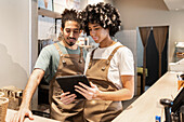 Two café employees in aprons work on a tablet behind a café counter to complete and plan tasks together The cosy ambience emphasises teamwork and the modern café culture
