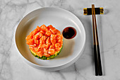 A gourmet dish with tender cubes of salmon on a bed of avocado, served with a drizzle of dark sauce on a white plate, accompanied by chopsticks