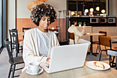 A young professional with vitiligo works on a laptop in a cosy café and enjoys a cup of coffee. The modern surroundings and natural light create a productive, relaxed atmosphere