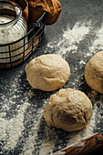 Fresh balls of dough lie on a black worktop dusted with flour. A glass jar filled with flour stands nearby, ready for the baking preparations. The scene captures a rustic kitchen atmosphere.