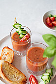 Zwei Gläser Gazpacho garniert mit Gurken- und Tomatenspießen, serviert mit geröstetem Brot Eine frische und farbenfrohe Präsentation, perfekt für eine leichte Mahlzeit