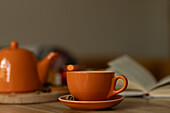A warm, inviting scene with an orange-coloured teacup and a teapot on a wooden table, accompanied by an open book in the background, creating the perfect relaxing atmosphere.