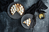 A delicious homemade lemon cake with toasted meringue, served on grey plates. Black cutlery and a glass with lemon slices complete the elegant table setting on a textured surface.